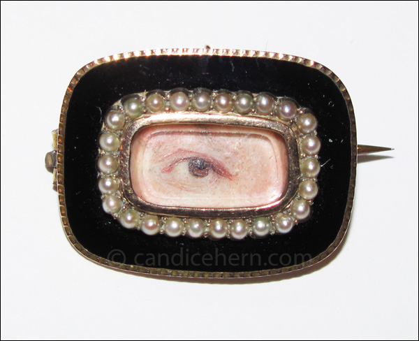 A gentleman’s brown eye painted in miniature on ivory, set in gold with black enamel and seed pearls. The combination of a black border and seed pearls (which can symbolize tears) marks this as a mourning brooch. ⅞” ⅝” Early 19th century.